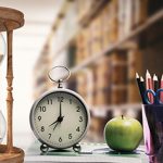 Hour glass, alarm clock, apple and pen holder against library in background